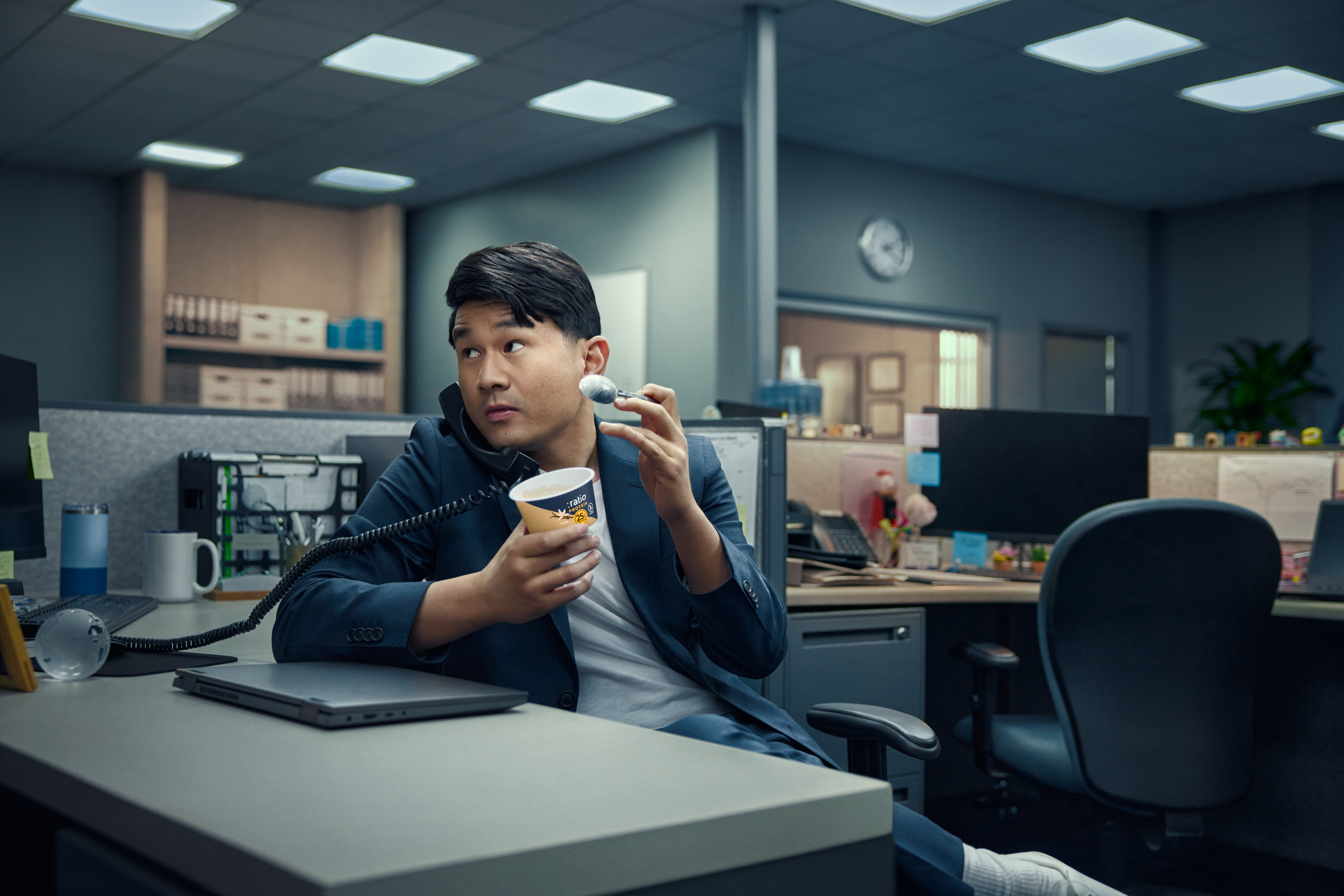 Comedian Ronny Chieng eating a :ratio dairy snack while sitting at an office cubical.